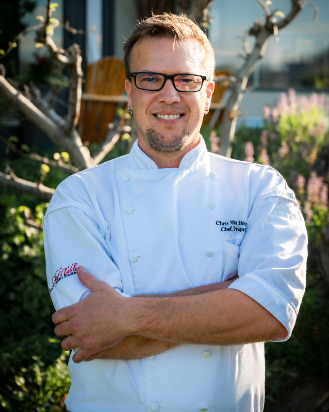 A photo of Chris Van Hooydonk standing in a white chef's jacket in front of a luscious garden of herbs posing for a photo, the esteemed chef at Backyard Farm Chef's Table.